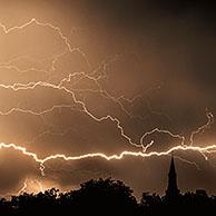 Rain falling and strokes of forked lightnings during thunderstorm at night over church tower and trees